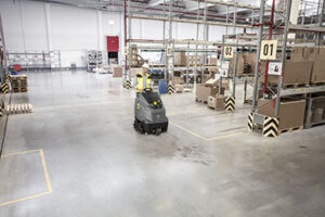 A warehouse floor-cleaning machine moving through an industrial storage area, representing industrial cleaning equipment used for managing hazardous spills and chemical spills. The scene relates to safely cleaning hazardous spills, using industrial wet/dry vacuums, steam cleaners for industrial floors, neutralising chemicals safely, and following a proper spill response plan with appropriate PPE for chemical spills.